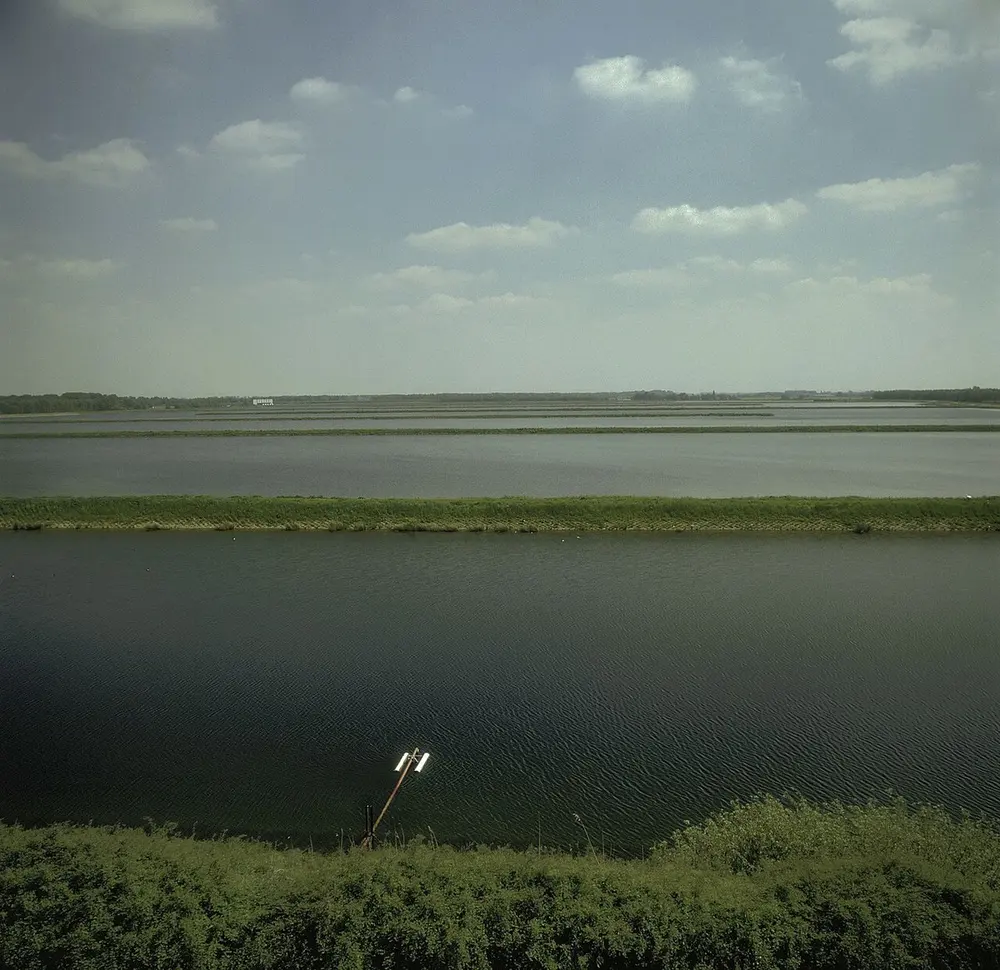 Brede, langgerekte waterbekkens achter elkaar, gescheiden door smalle begroeide dijken, met op de voorgrond struiken en een meetpaal in het water