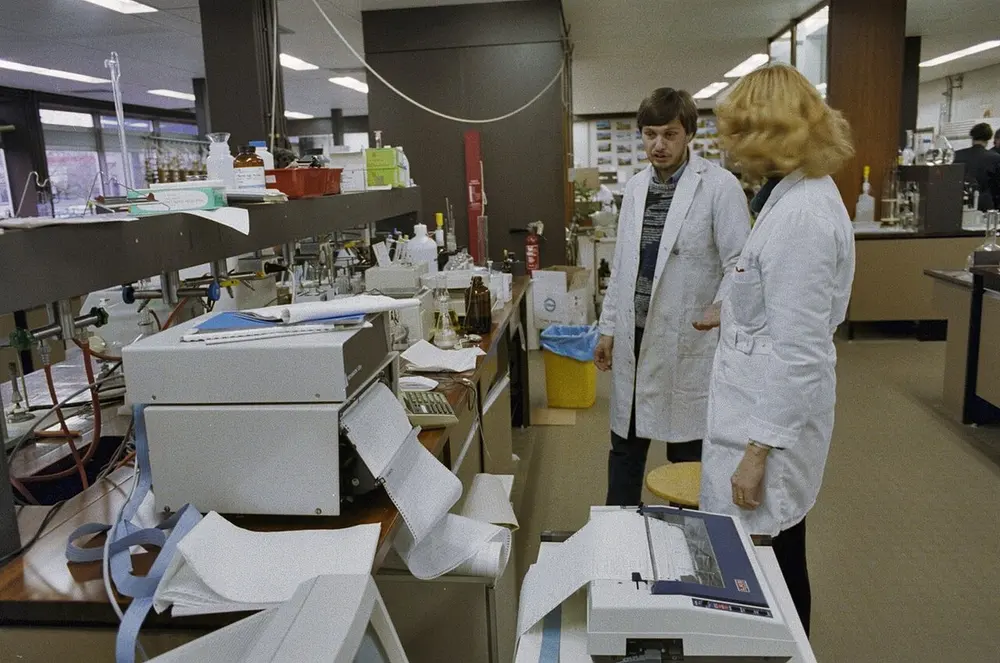 Laboratorium met werkbanken vol flessen en meetapparatuur; een man en een vrouw in witte jassen staan bij een printer die metingen op kettingpapier afdrukt