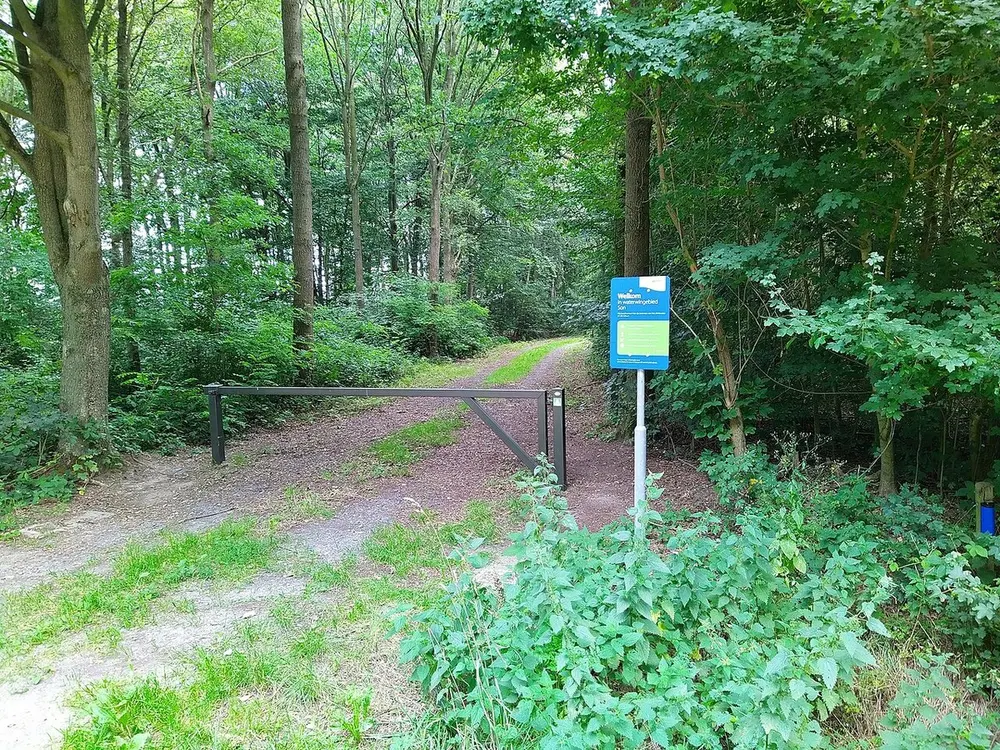 Bospad met een grijze slagboom; rechts een blauw bord met de tekst Welkom in waterwingebied Son, omringd door brandnetels en loofbomen