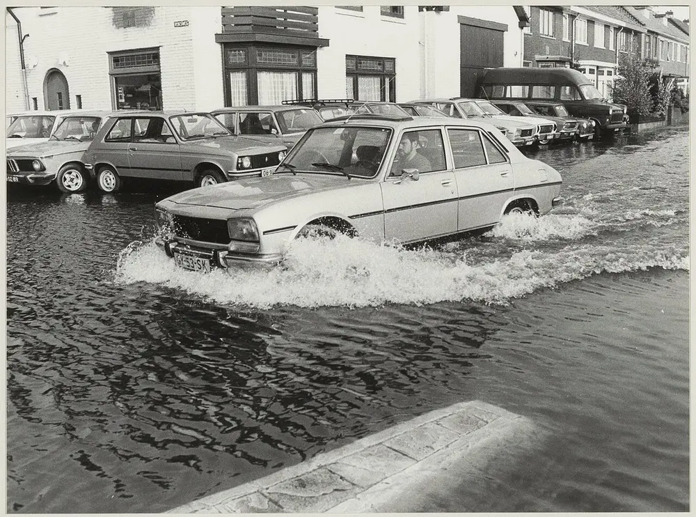 Zwart-witfoto van een auto die door een ondergelopen woonstraat rijdt en een golf water opzij duwt, met geparkeerde auto's langs de huizen