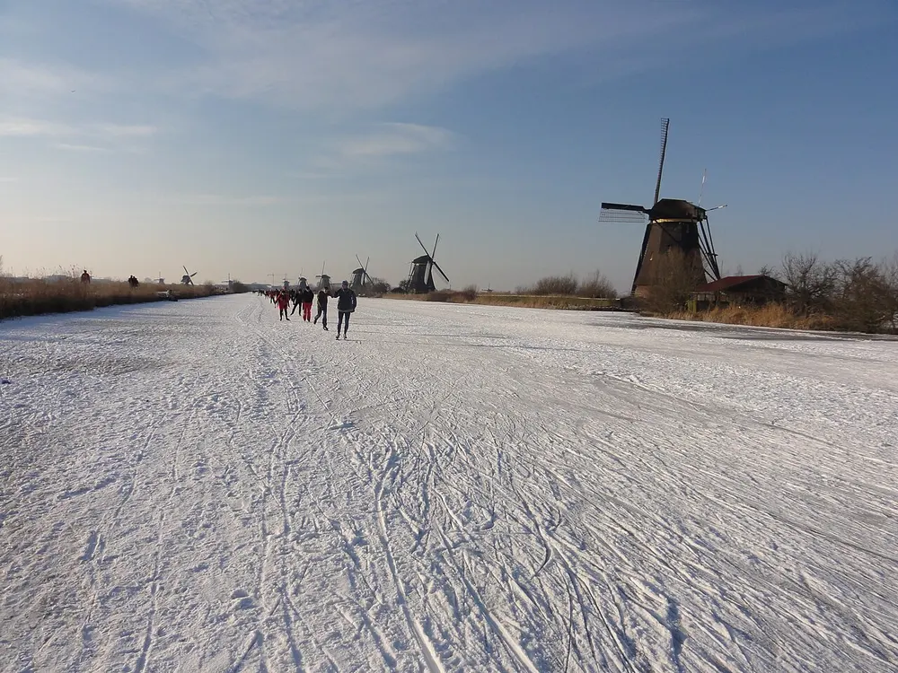 Tientallen schaatsers op een besneeuwde, bekraste ijsvlakte tussen rietkragen, met rechts een grote rietgedekte windmolen en in de verte een rij andere molens onder een blauwe lucht