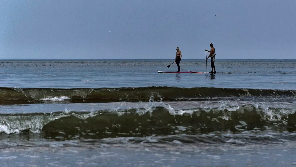 Twee mensen staand op lange supboards met een peddel in de hand, op kalme zee achter een rij brekende golven, onder een grijze lucht