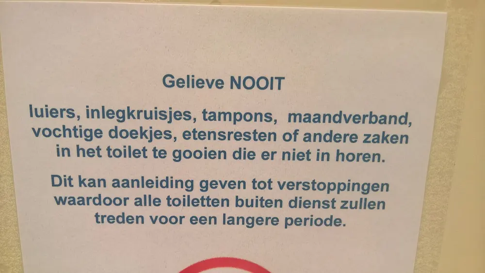 Wit papier aan een muur met blauwe tekst: gelieve nooit luiers, inlegkruisjes, tampons, maandverband, vochtige doekjes of etensresten in het toilet te gooien, met een waarschuwing voor verstoppingen