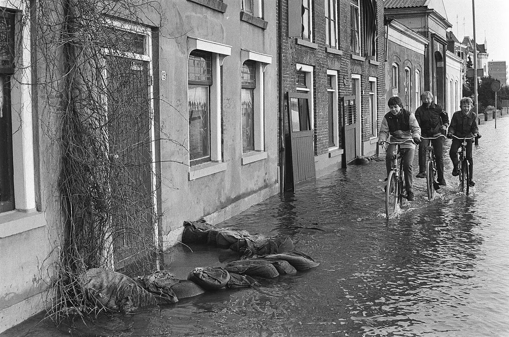 Zwart-witfoto van een ondergelopen straat met oude huizen, waar zandzakken tegen een voordeur en de gevel liggen en drie jongens door het water fietsen