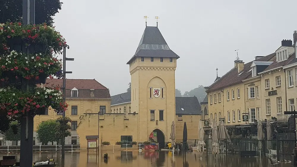 Plein in Valkenburg dat blank staat met bruin water, met op de achtergrond een lichtgele stadspoort met een leeuwenwapen en links hangende bloembakken aan een lantaarnpaal