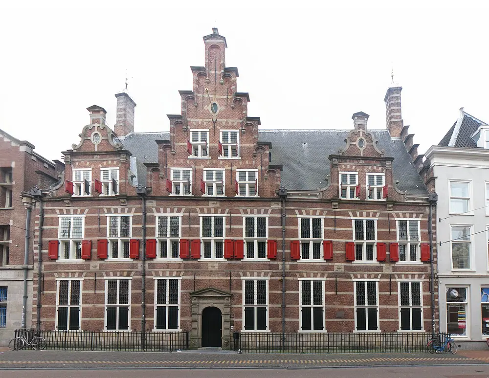 Gevel van een breed bakstenen gebouw met trapgevels, rode luiken naast de ramen en een stenen poortje als ingang, met fietsen tegen het hek op de stoep ervoor