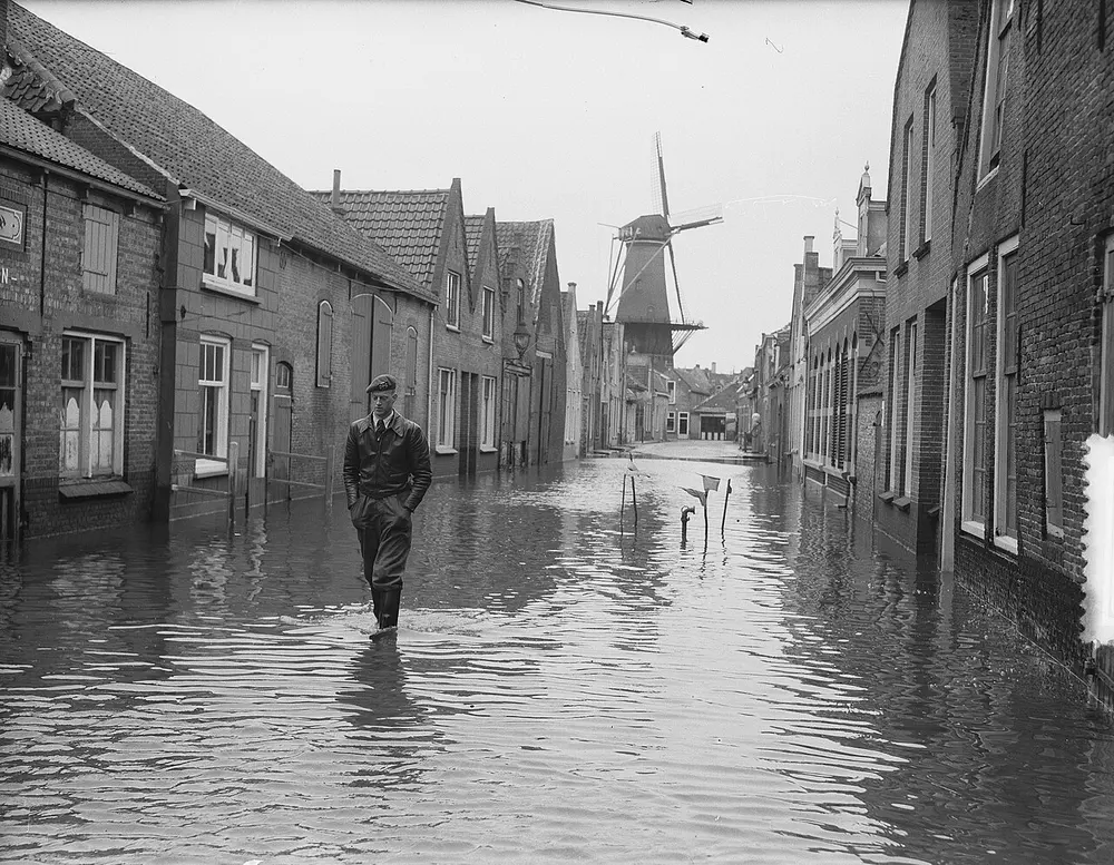 Zwart-witfoto van een man in laarzen die door een ondergelopen straat met lage bakstenen huizen loopt, met aan het eind van de straat een windmolen