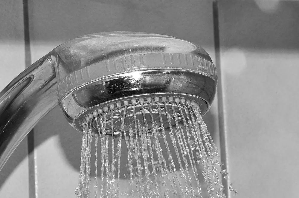 Zwart-witfoto van een verchroomde handdouche waaruit stralen water spuiten, tegen een betegelde wand