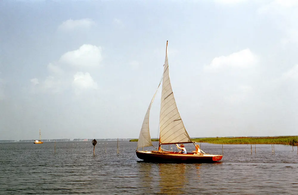 Kleurenfoto uit de jaren zestig van een open houten zeilboot met twee opvarenden op een breed meer met rietoevers, paaltjes in het water en in de verte een tweede zeilboot